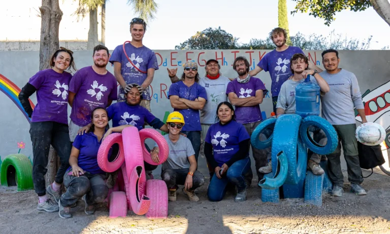 A group of 15 volunteers wearing purple and gray “All Hands and Hearts” shirts pose together outdoors in front of a painted wall featuring colorful letters and images. Some kneel beside large pink and blue elephant sculptures made from painted tires, while others stand smiling behind them.