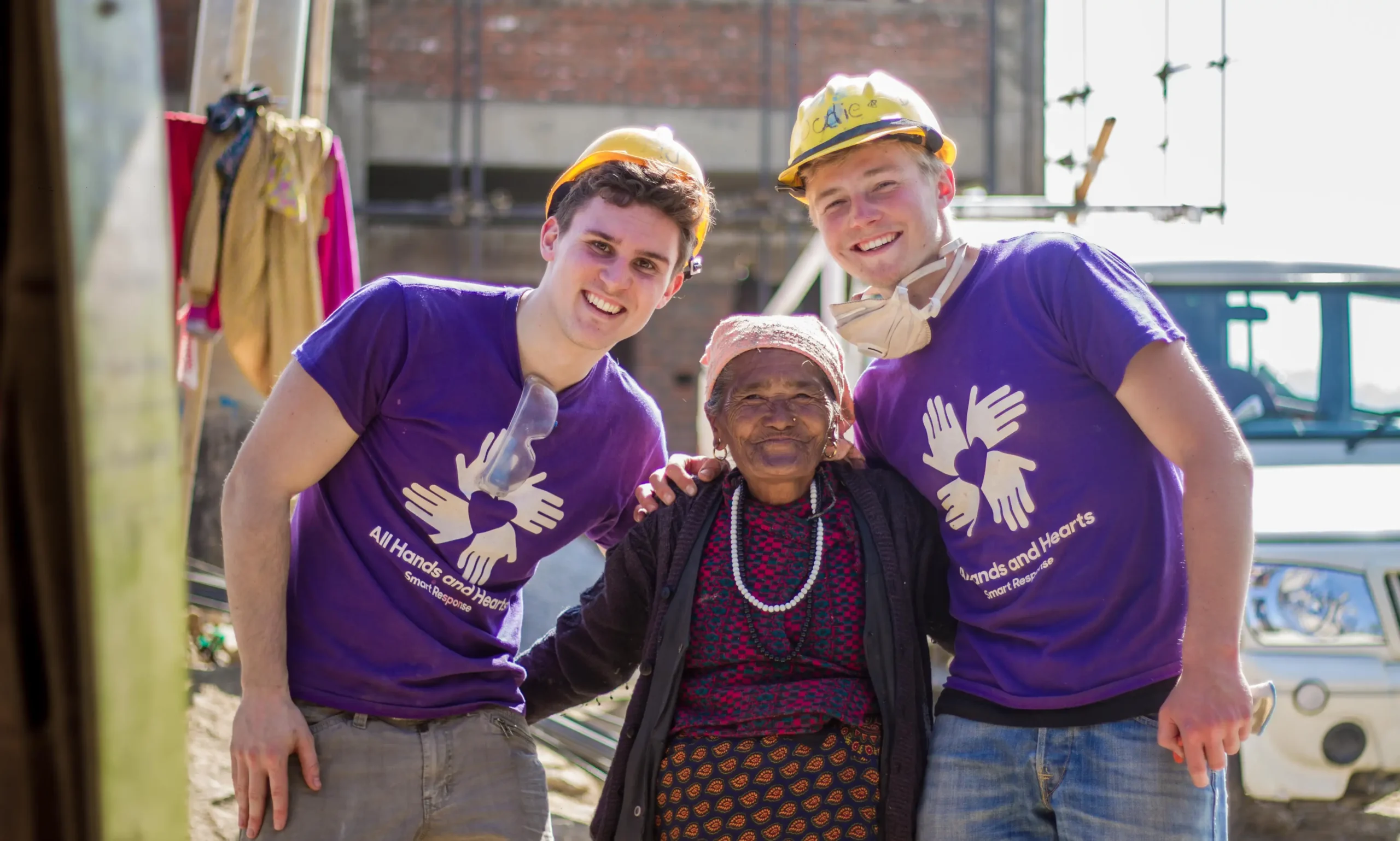Two young male volunteers in purple “All Hands and Hearts” shirts and yellow hard hats smile as they stand on either side of an older woman in traditional clothing. The trio poses closely together outside on a sunny day, with a white vehicle and construction area visible in the background.