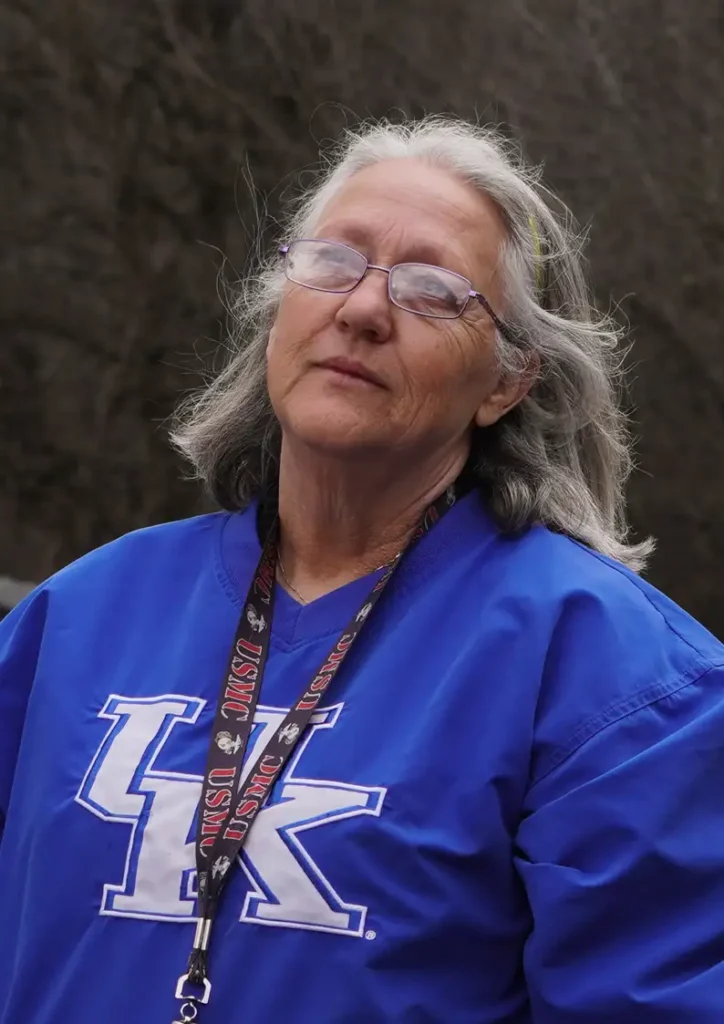 An older woman with gray hair and glasses stands outdoors wearing a bright blue University of Kentucky sweatshirt and a USMC lanyard. She looks calmly at the camera with a neutral expression, against a blurred natural background.
