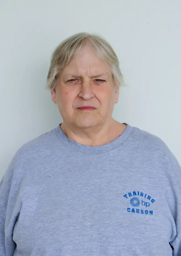 An older woman with short, light hair stands in front of a plain white background, wearing a light gray sweatshirt with a “Training BP Carson” logo printed in blue. She looks directly at the camera with a neutral expression.