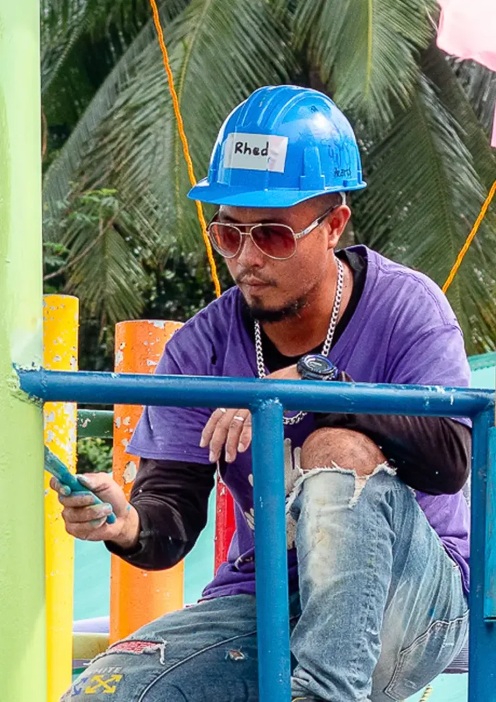 A man wearing a blue hard hat labeled "Rhed," sunglasses, and a purple volunteer shirt crouches on scaffolding while painting a green metal post. He appears focused on his work, surrounded by colorful poles and tropical palm trees in the background.