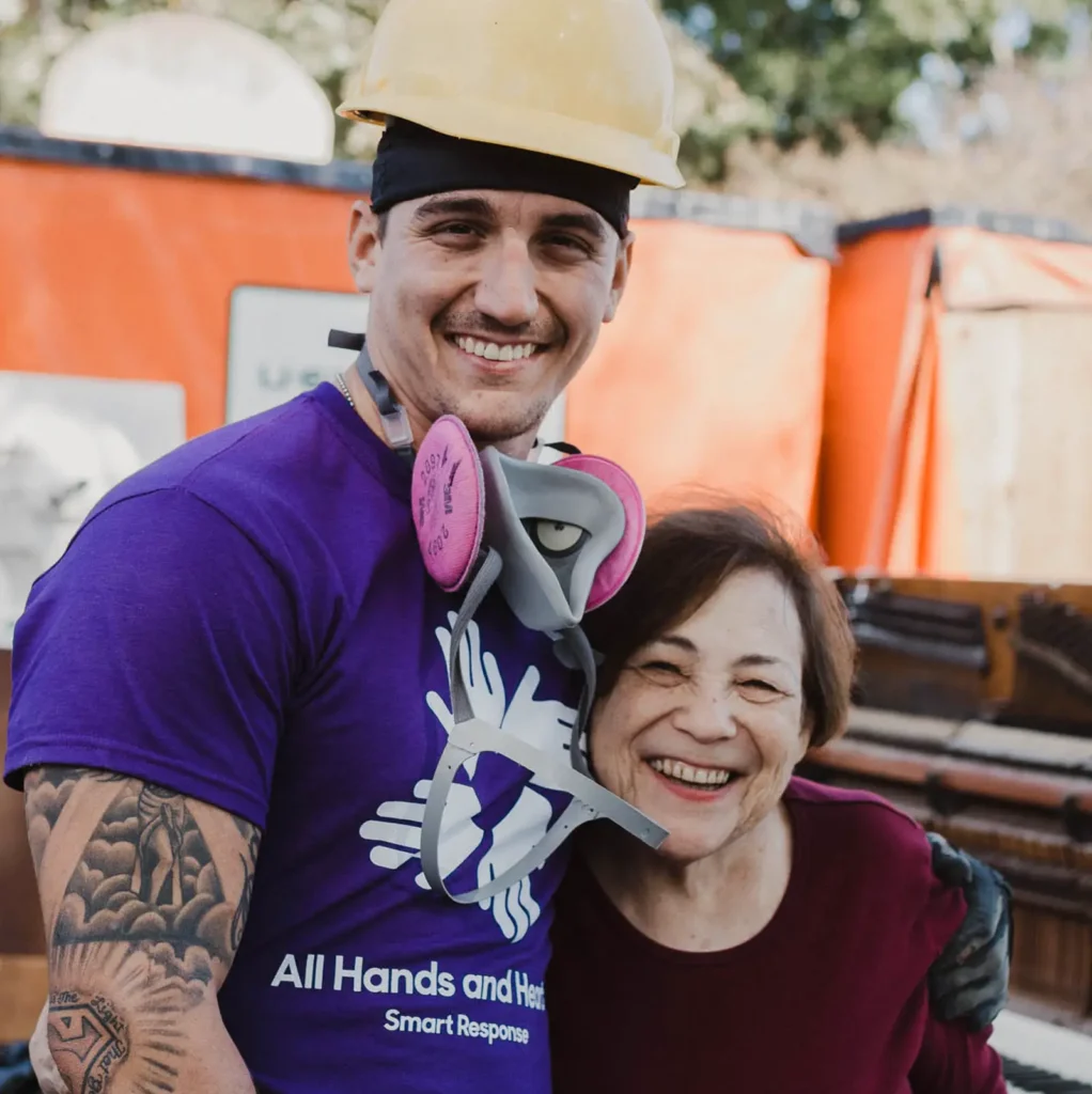 A smiling young male volunteer in a yellow hard hat and purple "All Hands & Hearts" shirt stands arm-in-arm with an elderly woman. They are outside near construction materials and bright orange tarps, radiating warmth and camaraderie.