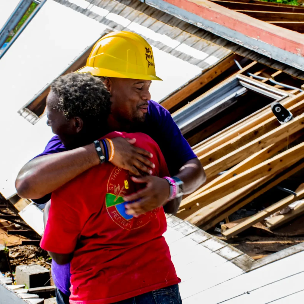 A volunteer in a yellow hard hat embraces a local community member in front of a pile of construction debris and wooden planks. Both individuals appear emotional, highlighting a moment of gratitude and connection during disaster relief efforts.