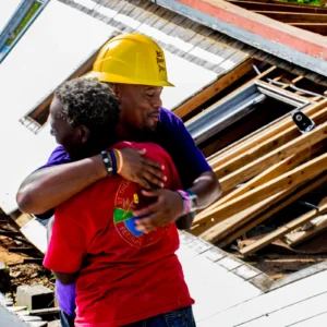 A volunteer in a yellow hard hat embraces a local community member in front of a pile of construction debris and wooden planks. Both individuals appear emotional, highlighting a moment of gratitude and connection during disaster relief efforts.