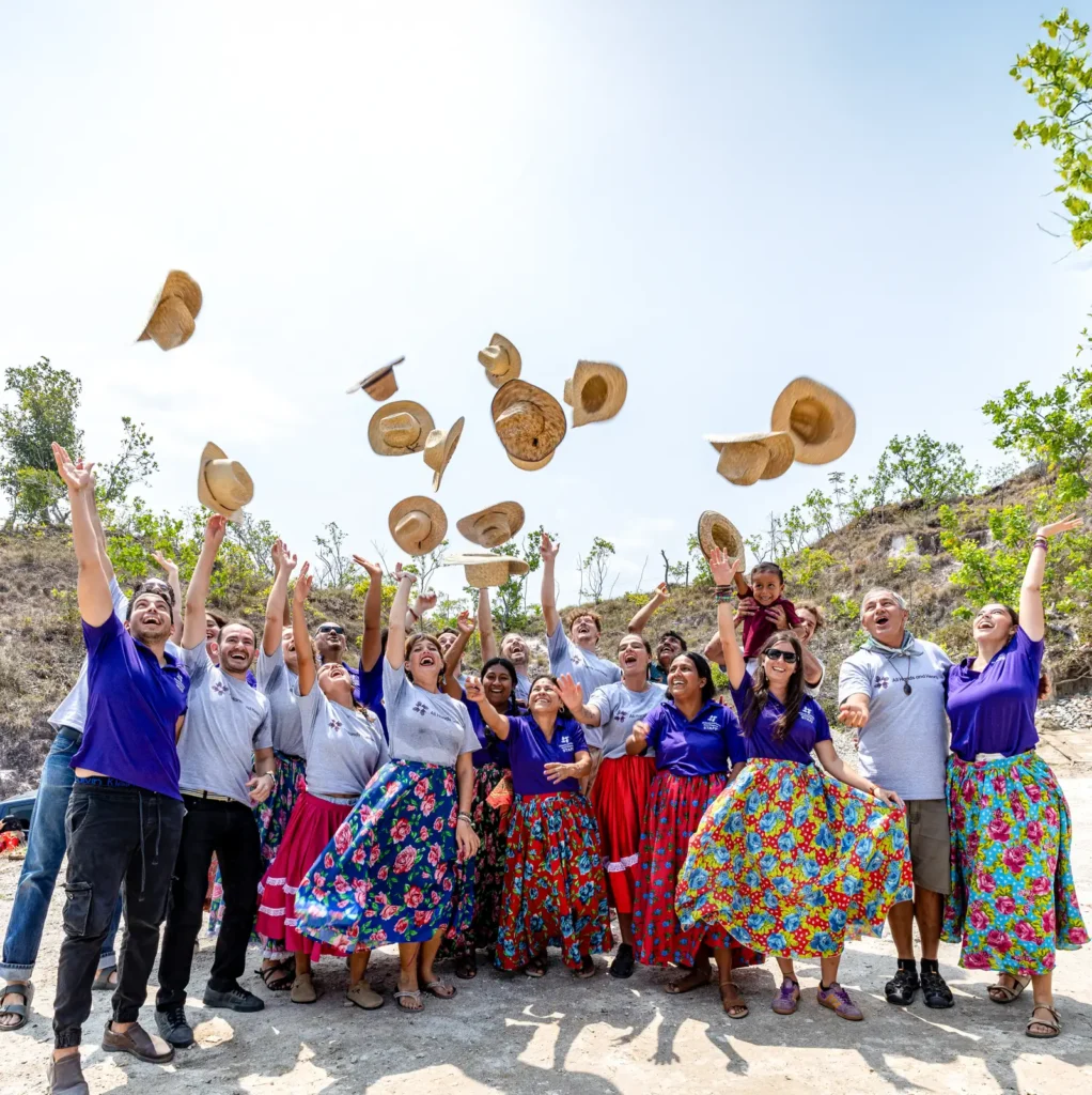 Volunteers and community members in colorful skirts and team shirts throwing straw hats in the air during a celebration outdoors.