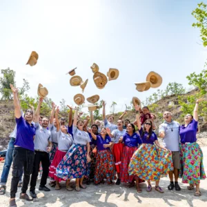 Volunteers and community members in colorful skirts and team shirts throwing straw hats in the air during a celebration outdoors.