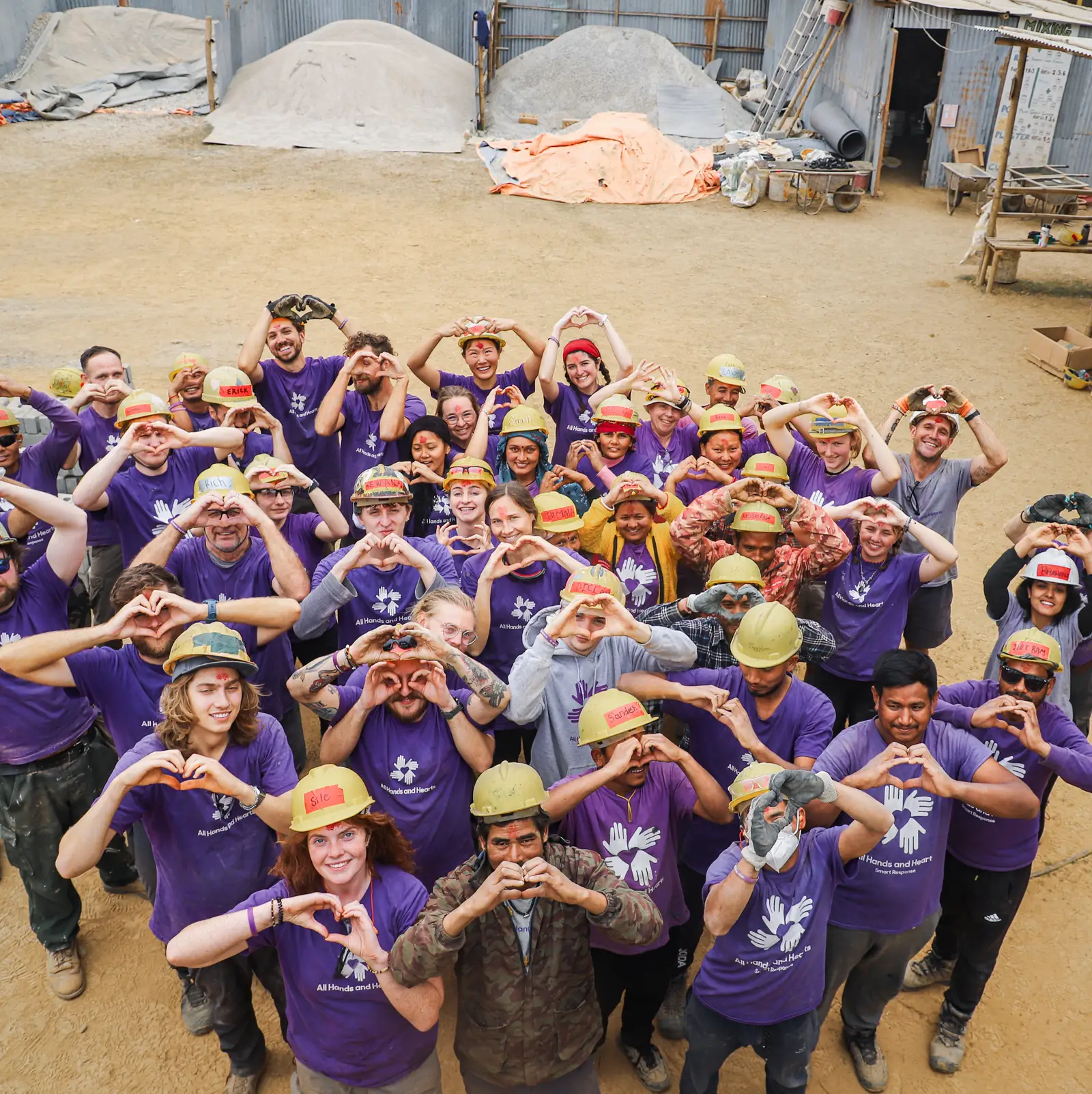 Large group of volunteers wearing purple shirts and hard hats smiling and making heart shapes with their hands.