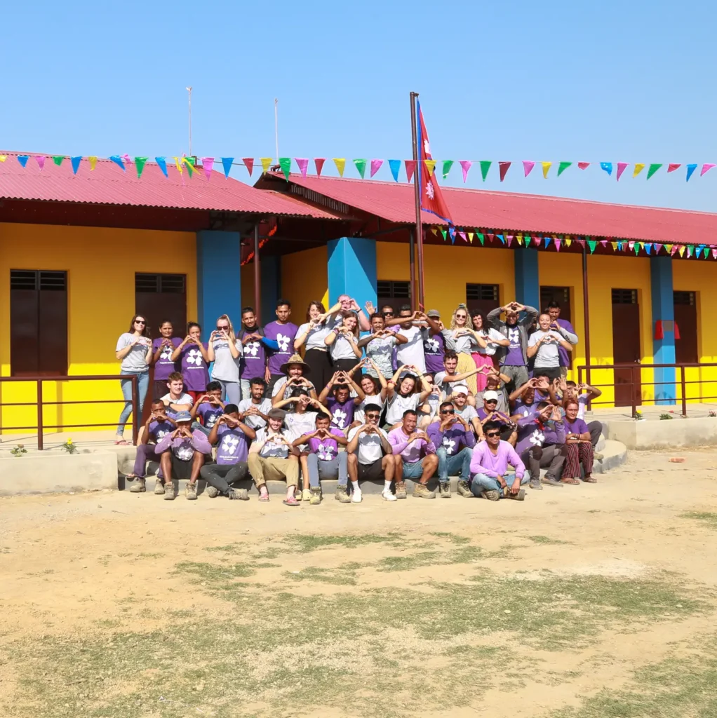 A large group of volunteers and community members pose happily in front of a newly completed school building painted in bright yellow, blue, and red. They are making heart shapes with their hands, and colorful pennant banners decorate the background under a clear blue sky.