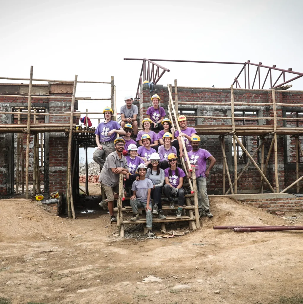 A group of volunteers in purple shirts and yellow hard hats pose together in front of a partially constructed brick building with scaffolding in Nepal. The group is smiling and appears to be taking a break from construction work.