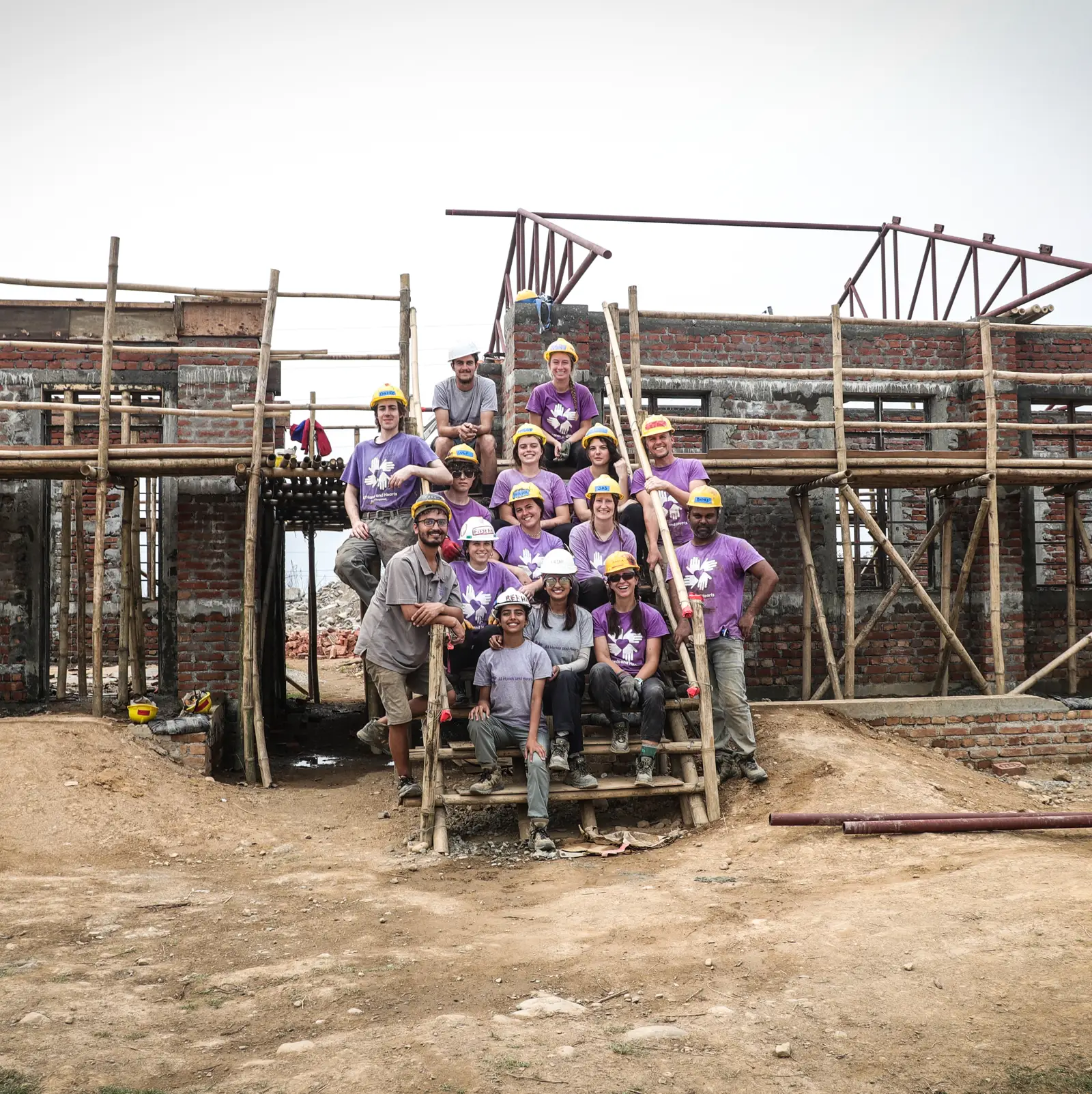 Un grupo de voluntarios con camisas moradas y cascos amarillos posan juntos delante de un edificio de ladrillo parcialmente construido con andamios en Nepal. El grupo sonríe y parece tomarse un descanso del trabajo de construcción.