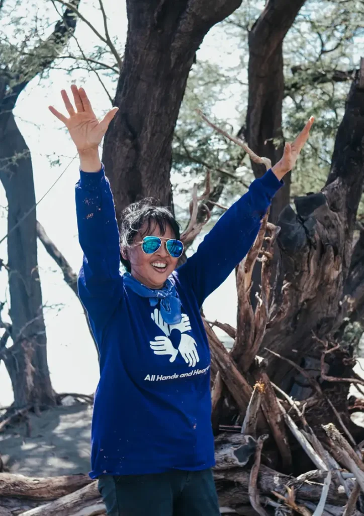 Woman wearing reflective sunglasses and a blue “All Hands and Hearts” shirt smiles with both arms raised in the air, standing in front of a forested area with tree trunks and driftwood.