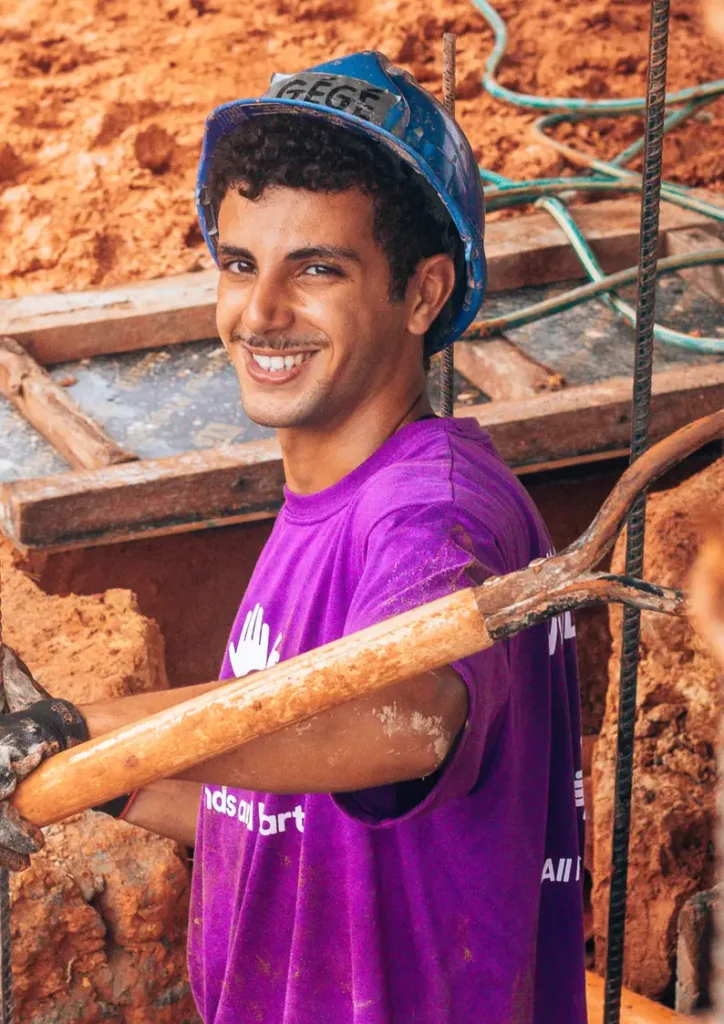 Young man wearing a blue hard hat labeled "GEBRIL" and a purple "All Hands and Hearts" shirt smiles at the camera while holding a construction tool at a muddy work site. Rebar and wooden framing are visible in the background.