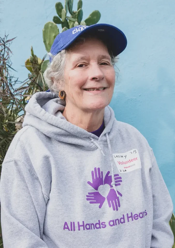Older woman smiling at the camera while wearing a blue cap and a gray hoodie with the “All Hands and Hearts” logo, along with a handwritten volunteer name tag reading “Lesley.” She stands in front of a light blue wall with a cactus in the background.