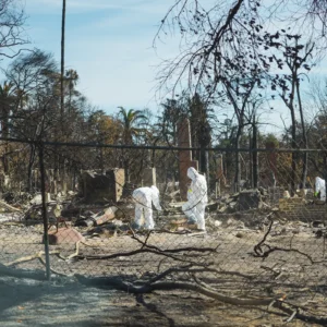 People in hazmat suits sift through debris and ash in a neighborhood destroyed by wildfire, with charred trees and remnants of buildings in the background.