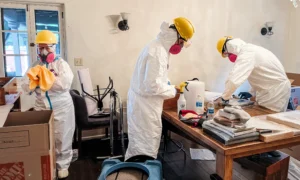 Tres voluntarios All Hands and Hearts con trajes de tyvek, cascos y mascarillas en el interior de una vivienda limpian enseres para remediar el humo.