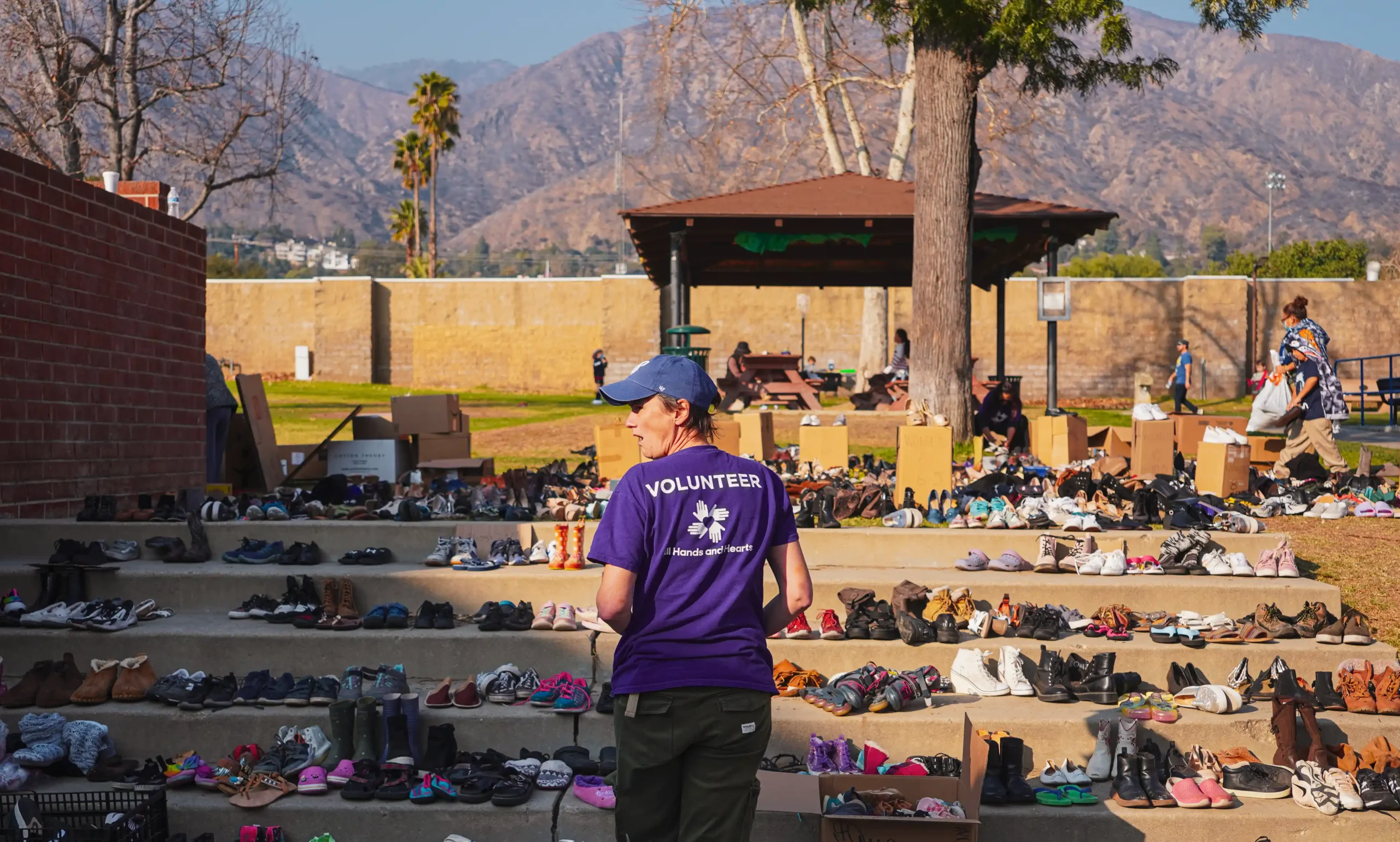 A volunteer wearing a purple "All Hands and Hearts" T-shirt stands among rows of donated shoes organized on outdoor steps, with boxes and other volunteers visible in the background, all set against a mountain backdrop.