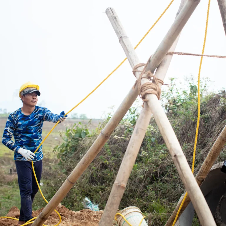 A construction worker in a yellow hard hat and blue camouflage shirt pulls a yellow rope attached to a wooden tripod structure, standing on a dirt mound in a rural outdoor setting with greenery in the background.