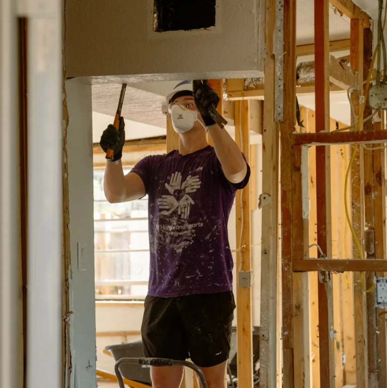 A volunteer in a protective mask, gloves, and a purple “All Hands and Hearts” shirt works inside a partially demolished house, using tools to remove drywall or ceiling materials amid exposed wooden framing.