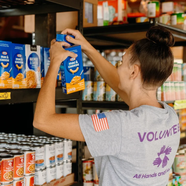 A volunteer wearing an “All Hands and Hearts” T-shirt organizes boxes of macaroni and cheese on a shelf in a food pantry, surrounded by rows of canned goods and other donations.