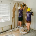 Two All Hands and Hearts volunteers inside a damaged home removing drywall