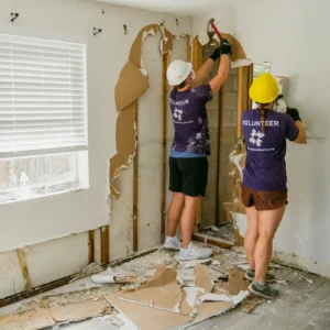 Two All Hands and Hearts volunteers inside a damaged home removing drywall
