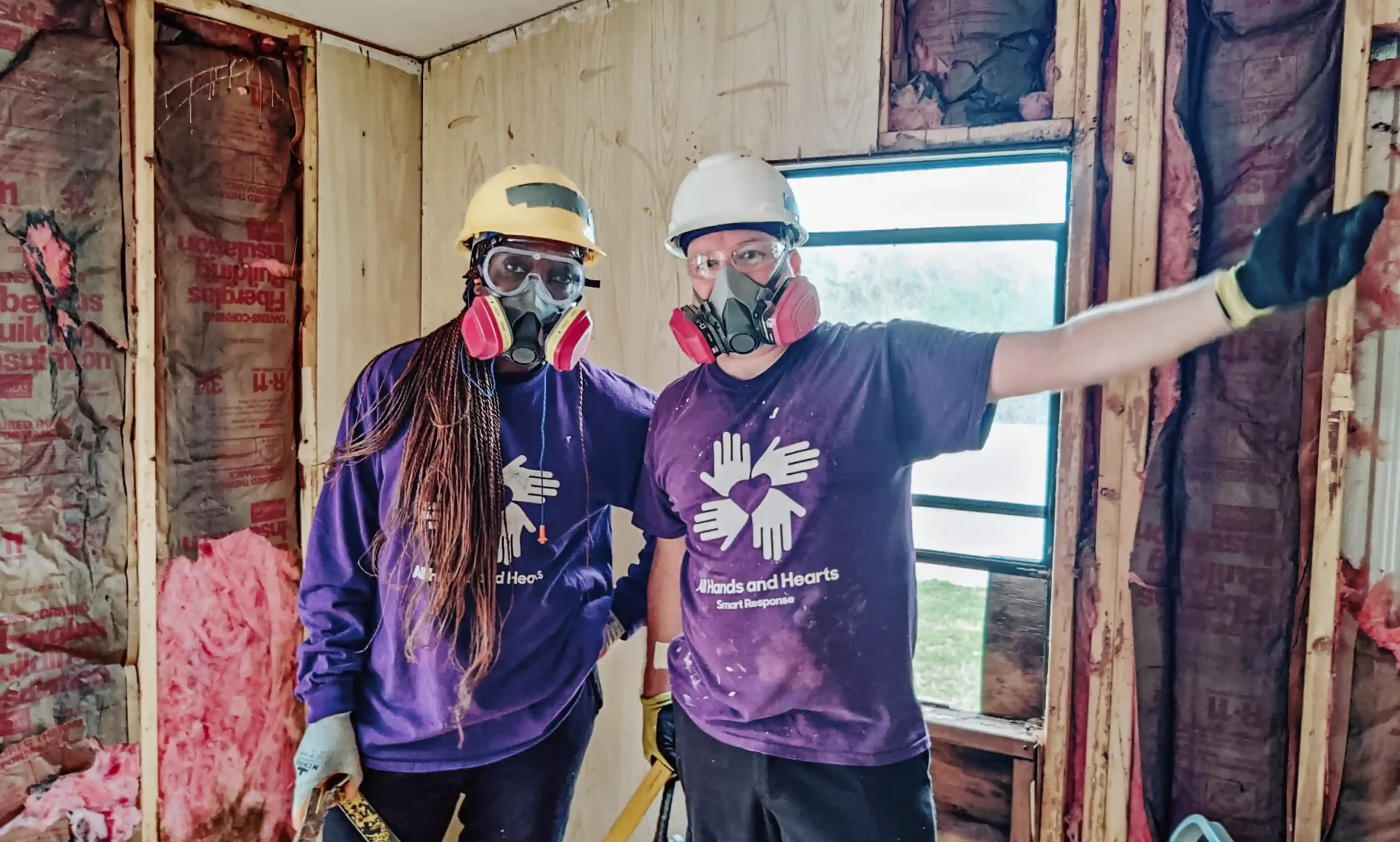 Two All Hands and Hearts volunteers in hard hats and respirators stand in a partially gutted home, with tools in hand