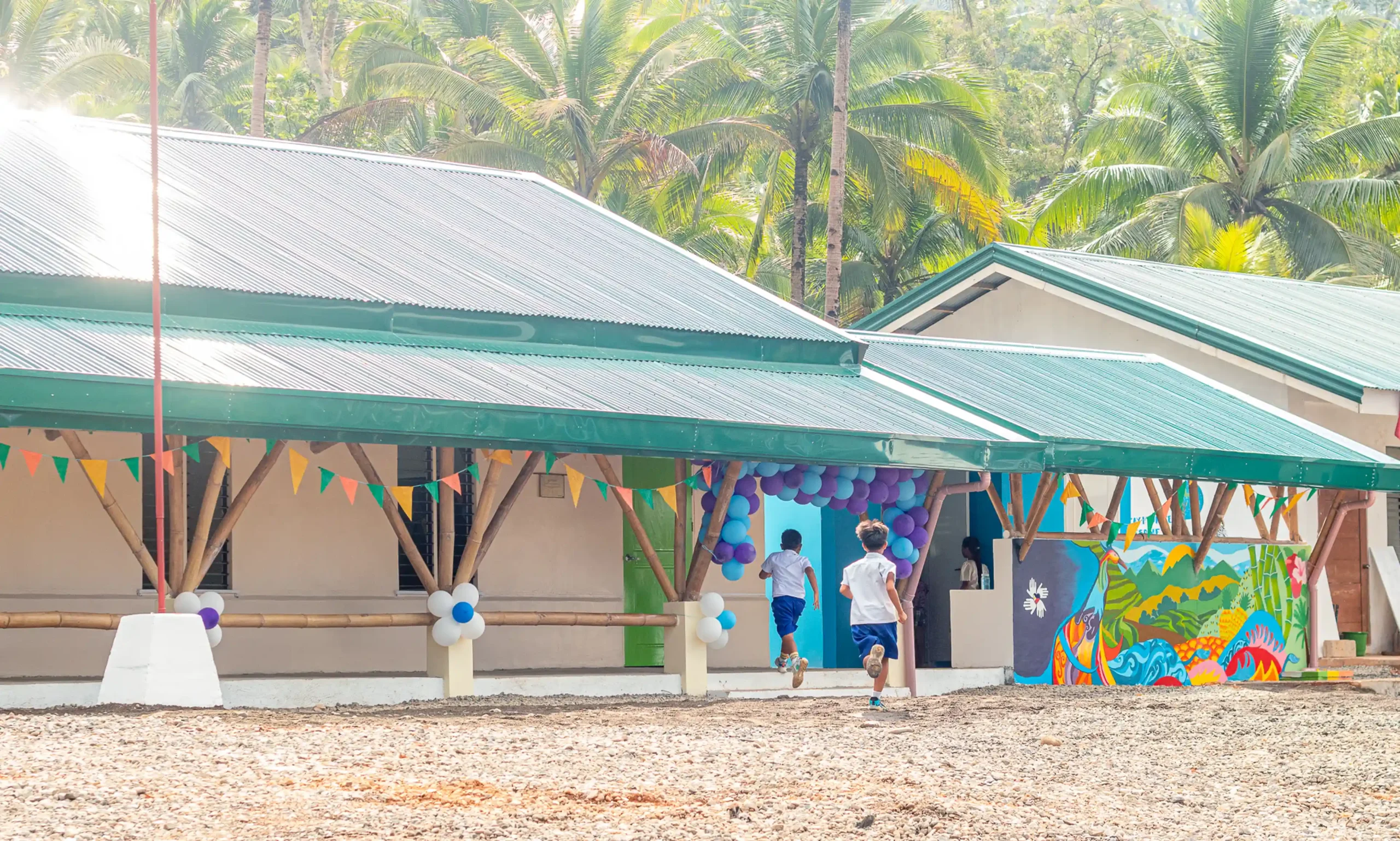 Two children run toward the entrance of a newly constructed school building, decorated with colorful balloons and festive flags. The school has a green metal roof, bamboo supports, and a vibrant mural on the wall. Palm trees fill the sunny background, creating a warm and tropical atmosphere.