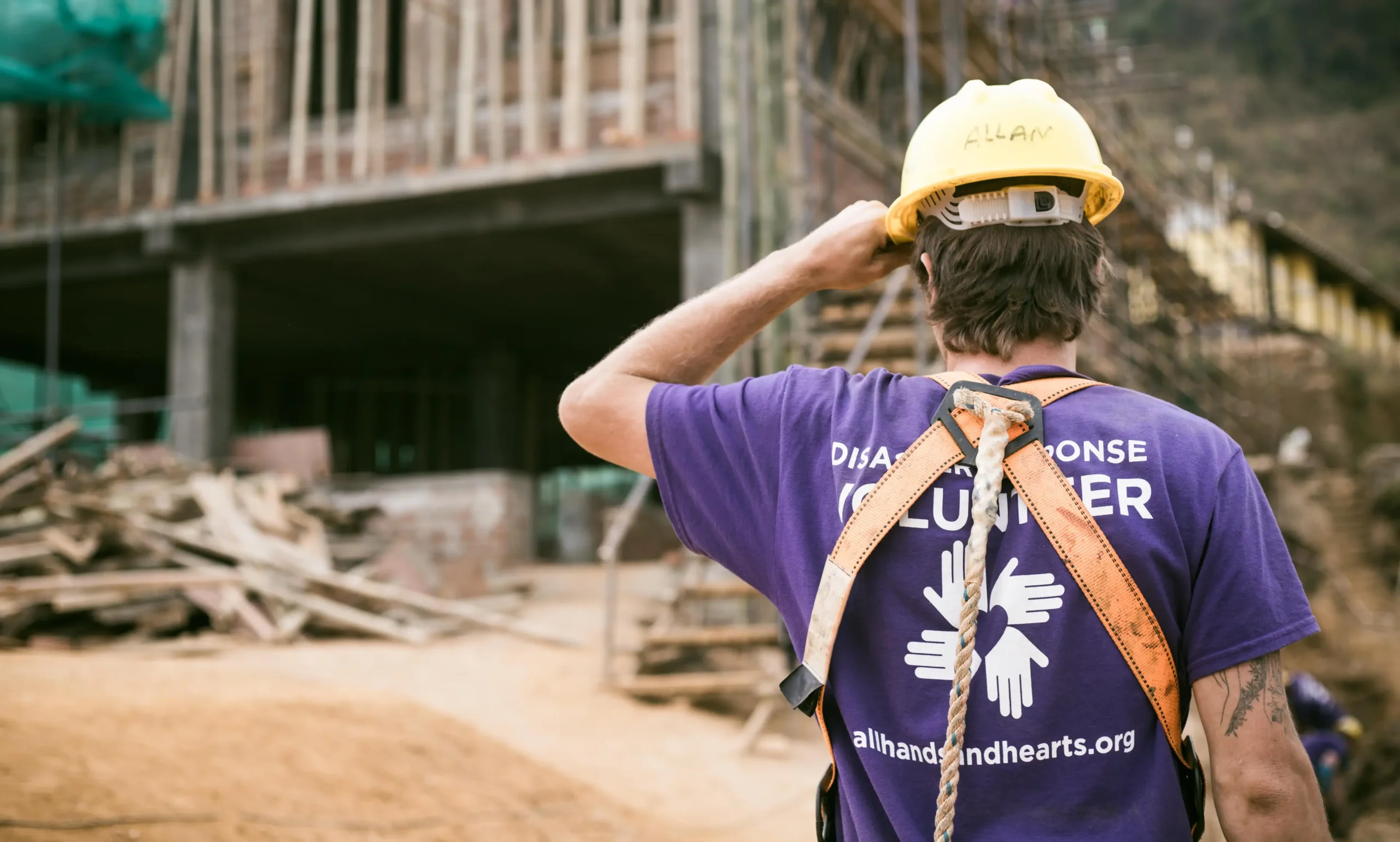 A disaster response volunteer wearing a safety harness and yellow hard hat stands with his back to the camera, looking toward a large construction site. He wears a purple shirt with the "All Hands and Hearts" logo and website on the back.