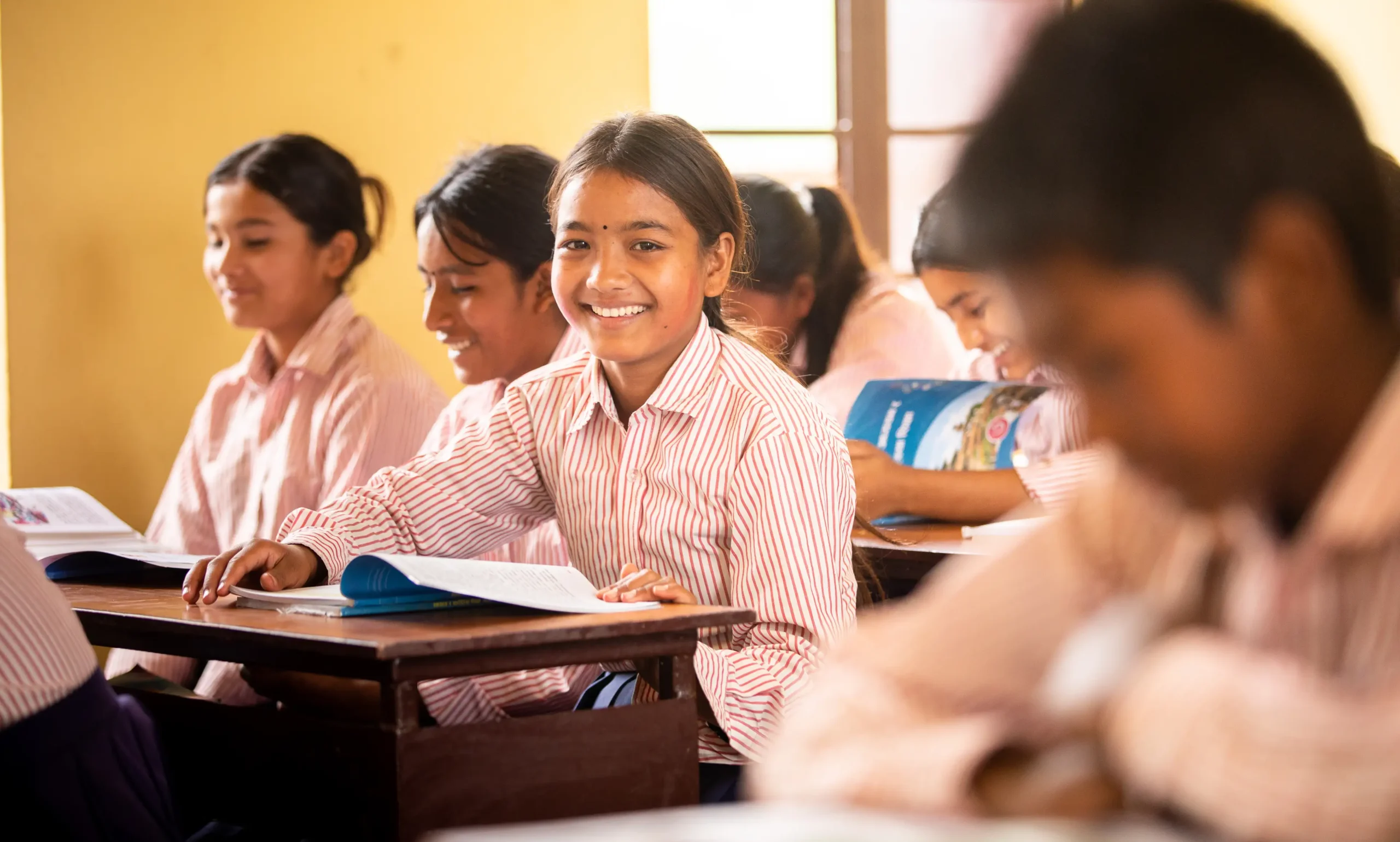 A group of schoolchildren in Nepal, wearing striped uniforms, sit at desks in a brightly lit classroom. One girl in the center smiles directly at the camera while holding an open book, surrounded by classmates who are reading and laughing.