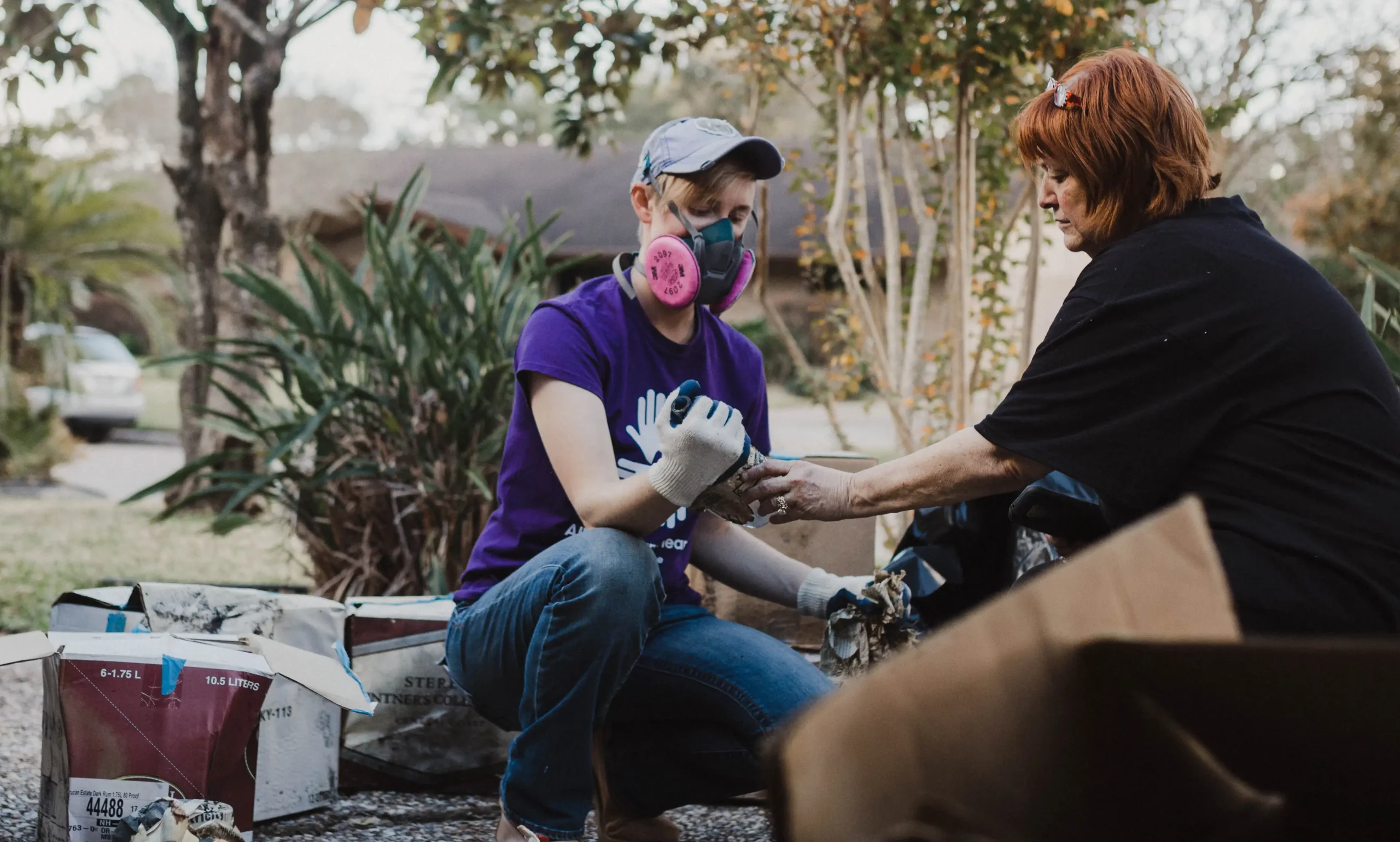 A volunteer wearing a purple “All Hands and Hearts” shirt, gloves, and a respirator mask kneels while helping an older woman sort through damaged belongings outside a home, surrounded by debris and greenery.