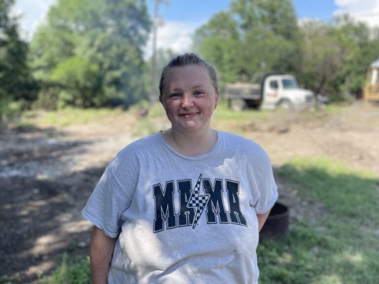 A young woman in a light gray MMA t-shirt smiles at the camera while standing at a disaster relief site, with a truck and trees in the background.