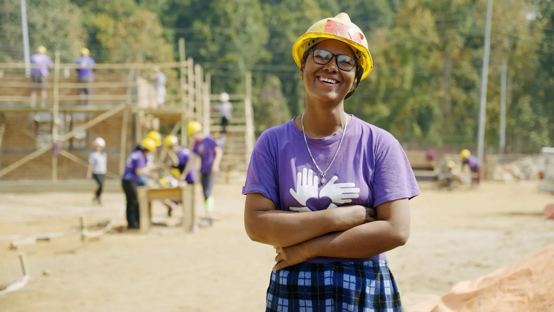 A smiling volunteer in a yellow hard hat and purple “All Hands and Hearts” shirt stands confidently at a construction site, with a partially built structure and fellow volunteers working in the background.
