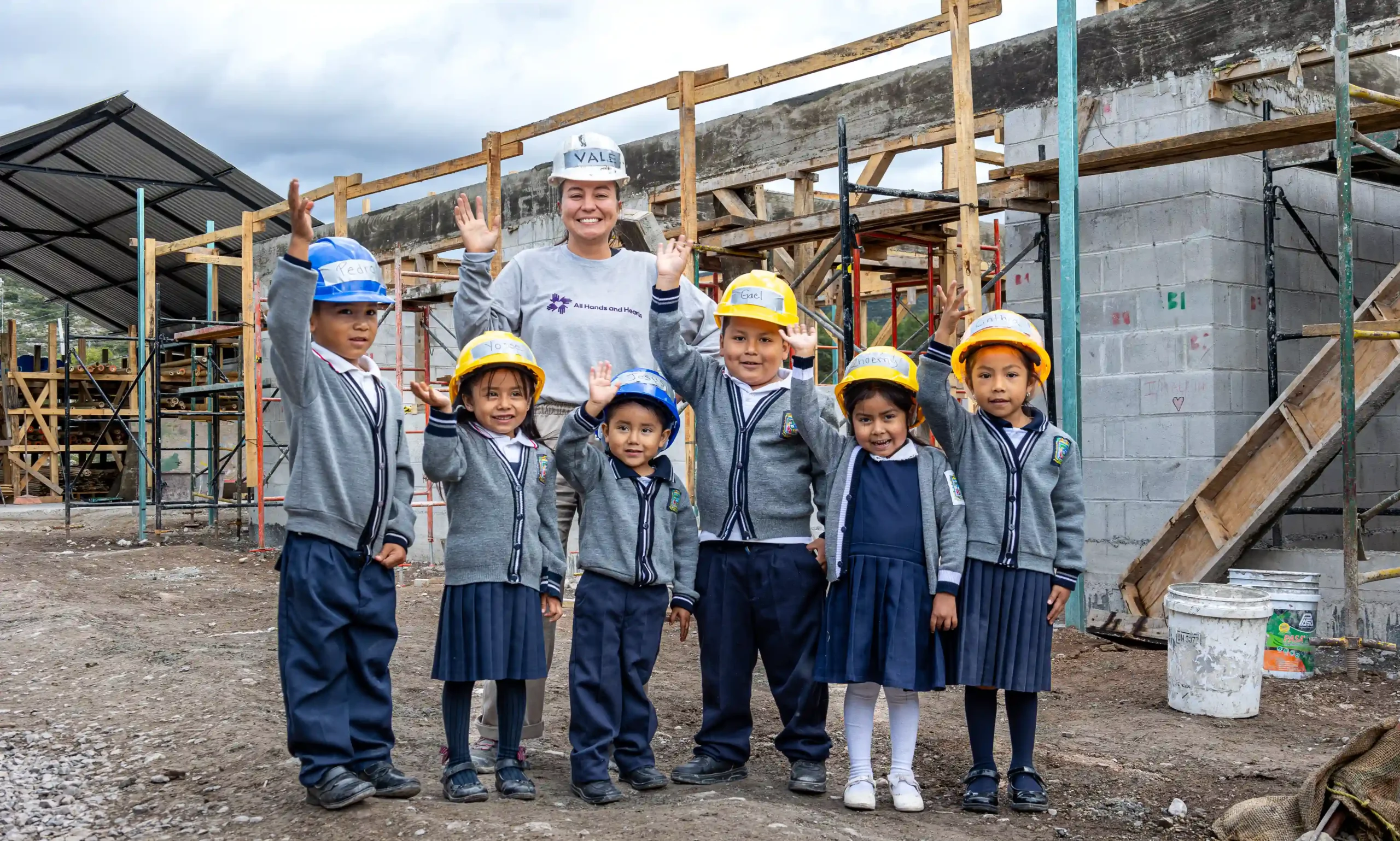 En las obras de una escuela, un grupo de niños con uniforme escolar y casco, junto con un miembro del personal All Hands and Hearts , saludan y sonríen.