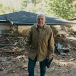 A homeowner stands in front of his severely damaged home; the home is barely visible behind a huge pile of fallen trees