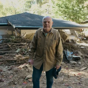 A homeowner stands in front of his severely damaged home; the home is barely visible behind a huge pile of fallen trees