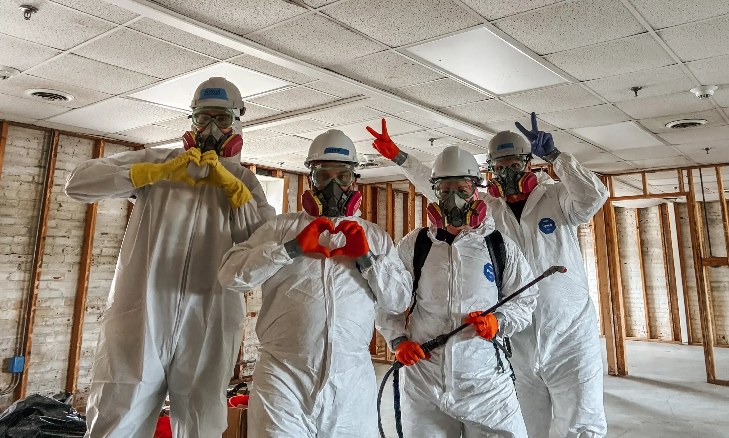 A group of All Hands and Hearts volunteers wearing Tyvek suits, hard hats and respirators pose in a gutted home holding mold sanitation equipment