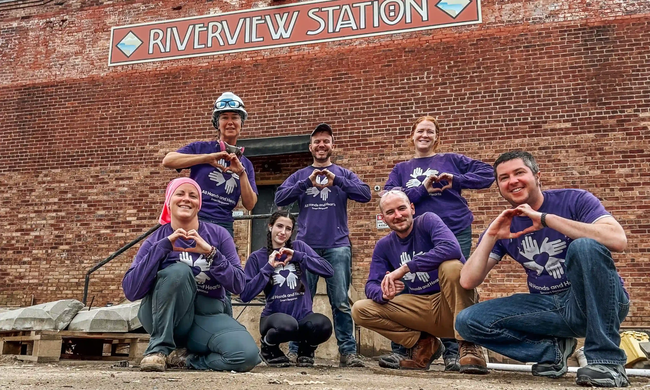 A group of All Hands and Hearts volunteers pose in front of Riverview Station making heart symbols with their hands