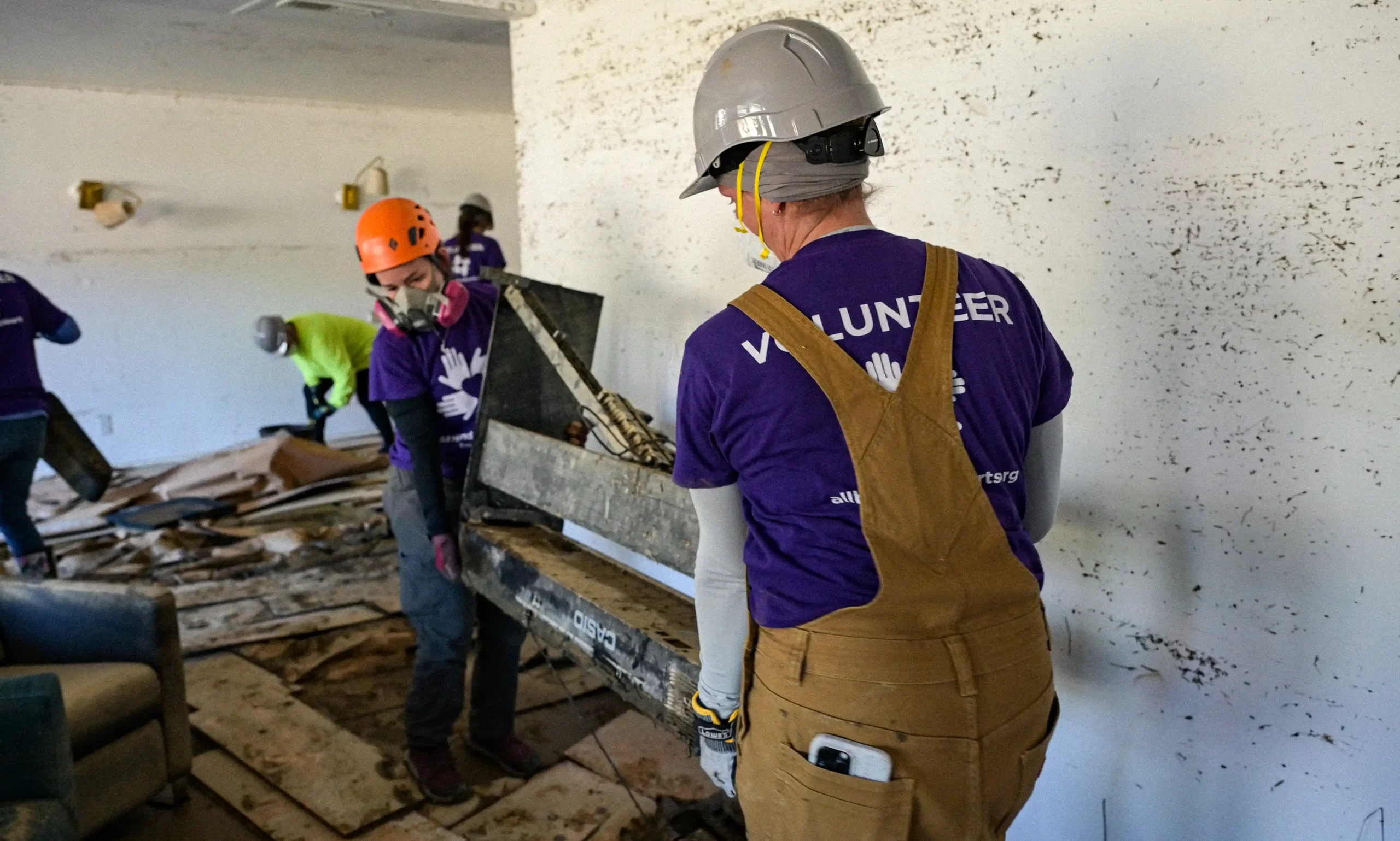 Two All Hands and Hearts volunteers wearing hard hats and respirators carry a work bench inside a flood damaged home