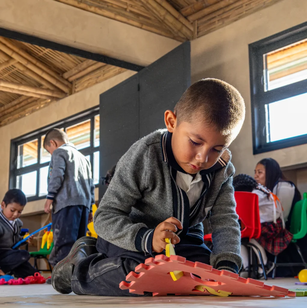 A young boy in a gray school uniform kneels on the floor of a brightly lit classroom, focused on assembling a colorful foam puzzle. Other children are engaged in similar activities in the background, surrounded by red and green chairs and educational toys. Sunlight streams in through the windows, illuminating the warm, wooden interior.