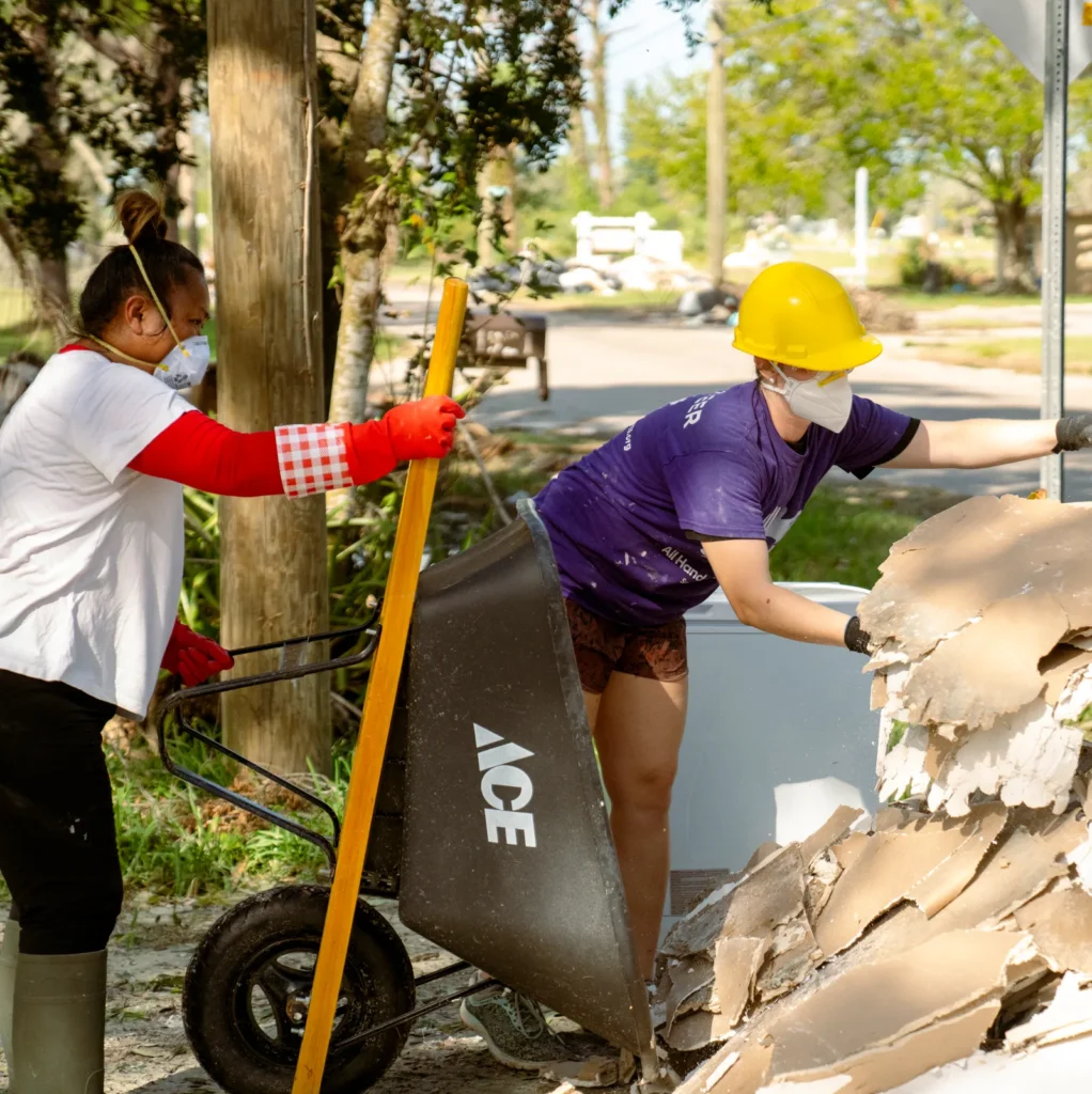 Two volunteers wearing protective masks and gloves clear debris into a wheelbarrow in a residential neighborhood. One person wears a yellow hard hat and a purple "All Hands and Hearts" shirt, while the other assists with a broom and stands nearby. Sunlight filters through the trees as they work amidst hurricane damage.