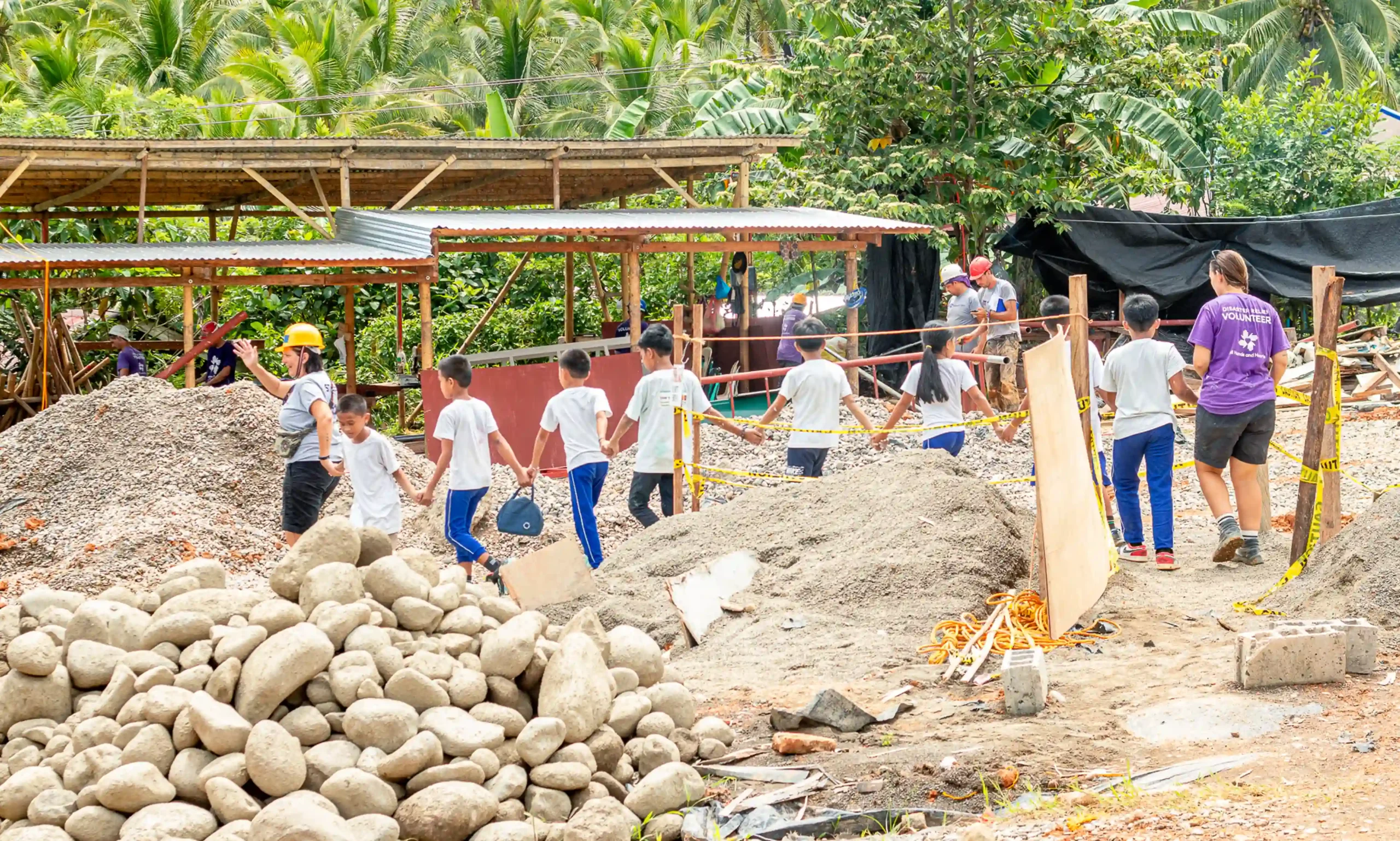 Una fila de estudiantes cogidos de la mano con un miembro del personal All Hands and Hearts al frente y un voluntario al fondo caminan por un centro escolar.