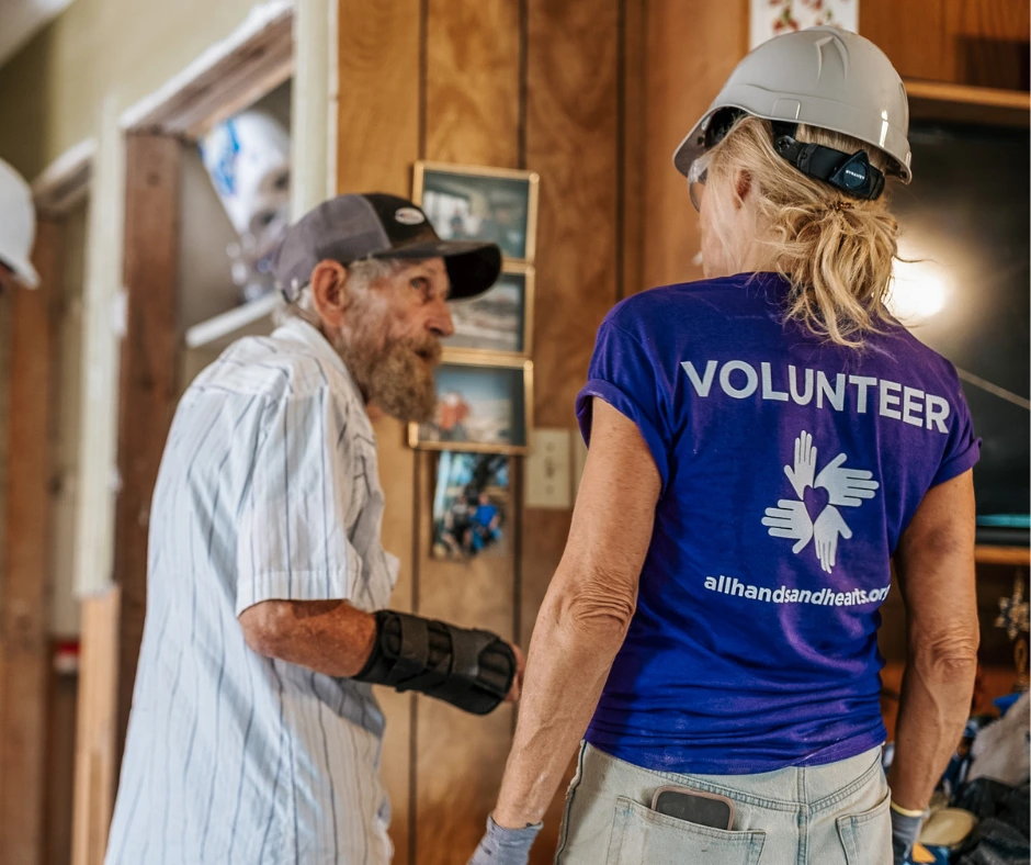 Una voluntaria con una camiseta morada deAll Hands and Hearts&quot; y un casco blanco habla con un anciano dentro de su casa, apoyando las labores de recuperación tras la catástrofe.
