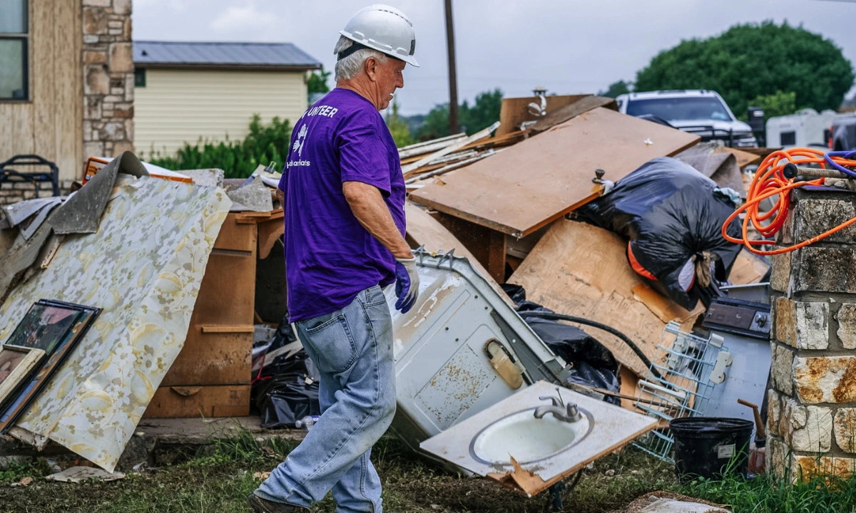 Un voluntario con casco y camiseta morada deAll Hands and Hearts retira los escombros de una casa tras una catástrofe natural, con las pertenencias dañadas apiladas tras él.