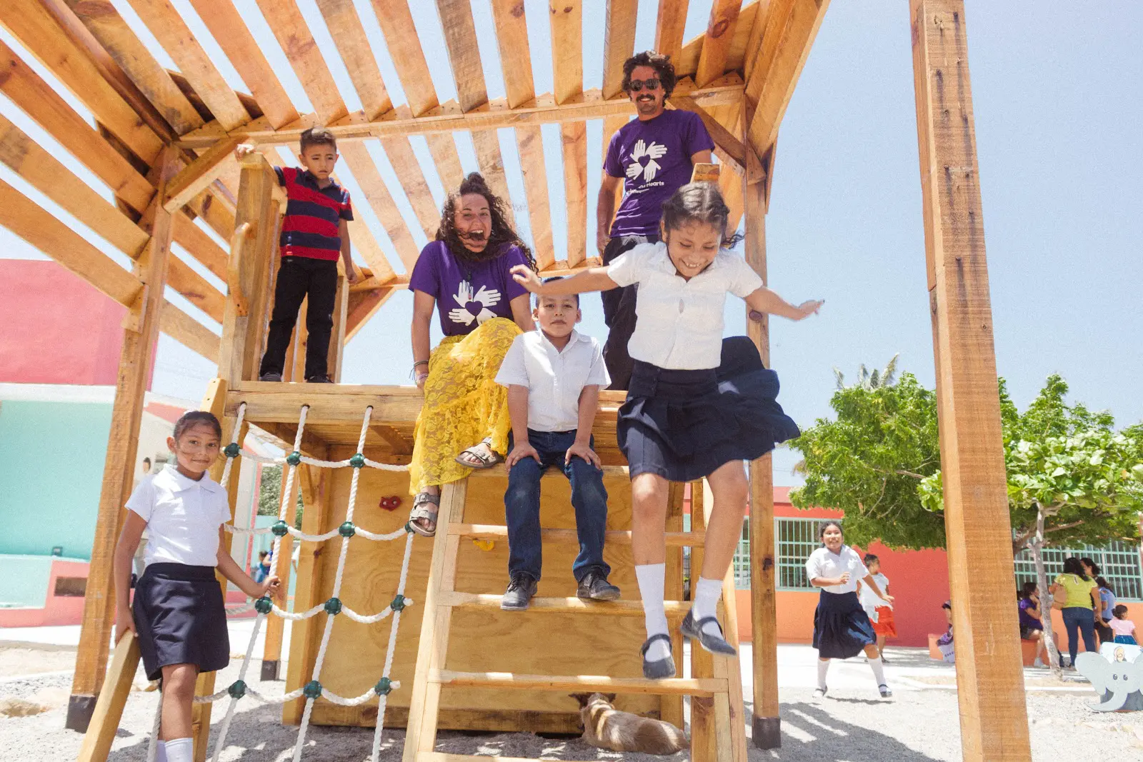 Children in school uniforms play on a wooden playground structure, while volunteers in purple All Hands and Hearts shirts stand nearby, supervising and sharing in the joyful moment.