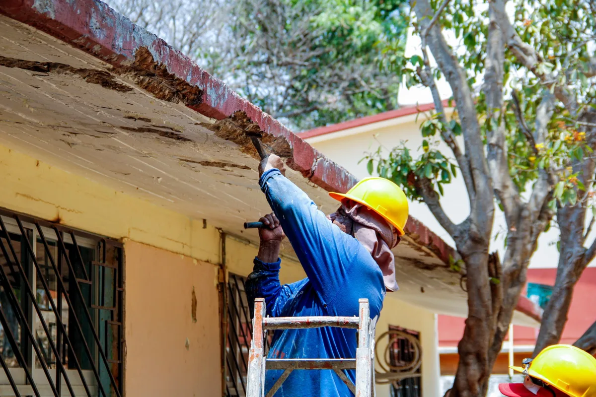 Volunteer in a yellow hard hat repairing a hurricane-damaged roof with a hammer and chisel.