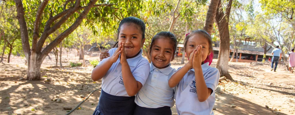 Three young girls in school uniforms smiling outdoors at a community school impacted by the hurricane.