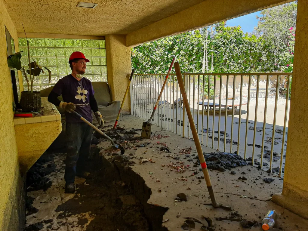 A volunteer in a hard hat and purple All Hands and Hearts shirt uses a shovel to clear thick mud and debris from a covered patio after flooding, with additional shovels leaning against the railing.