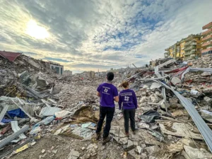 Two people in purple All Hands and Hearts shirts stand facing massive piles of rubble and collapsed buildings after the Turkey earthquake, with the sky breaking through above the devastation.