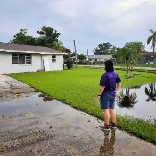 A person in a purple shirt stands on a driveway, observing waterlogged grass and floodwater pooling around a single-story home.
