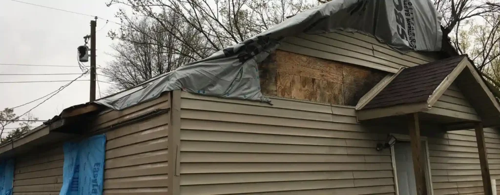 A home with part of the roof missing, covered by a blue tarp, showing severe tornado damage.